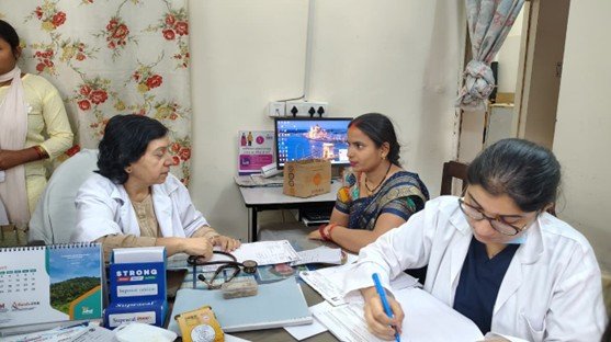 Three medical professionals sit at a desk in a clinic, one writing while another discusses with a patient in a sari; medical paperwork and a computer are visible.