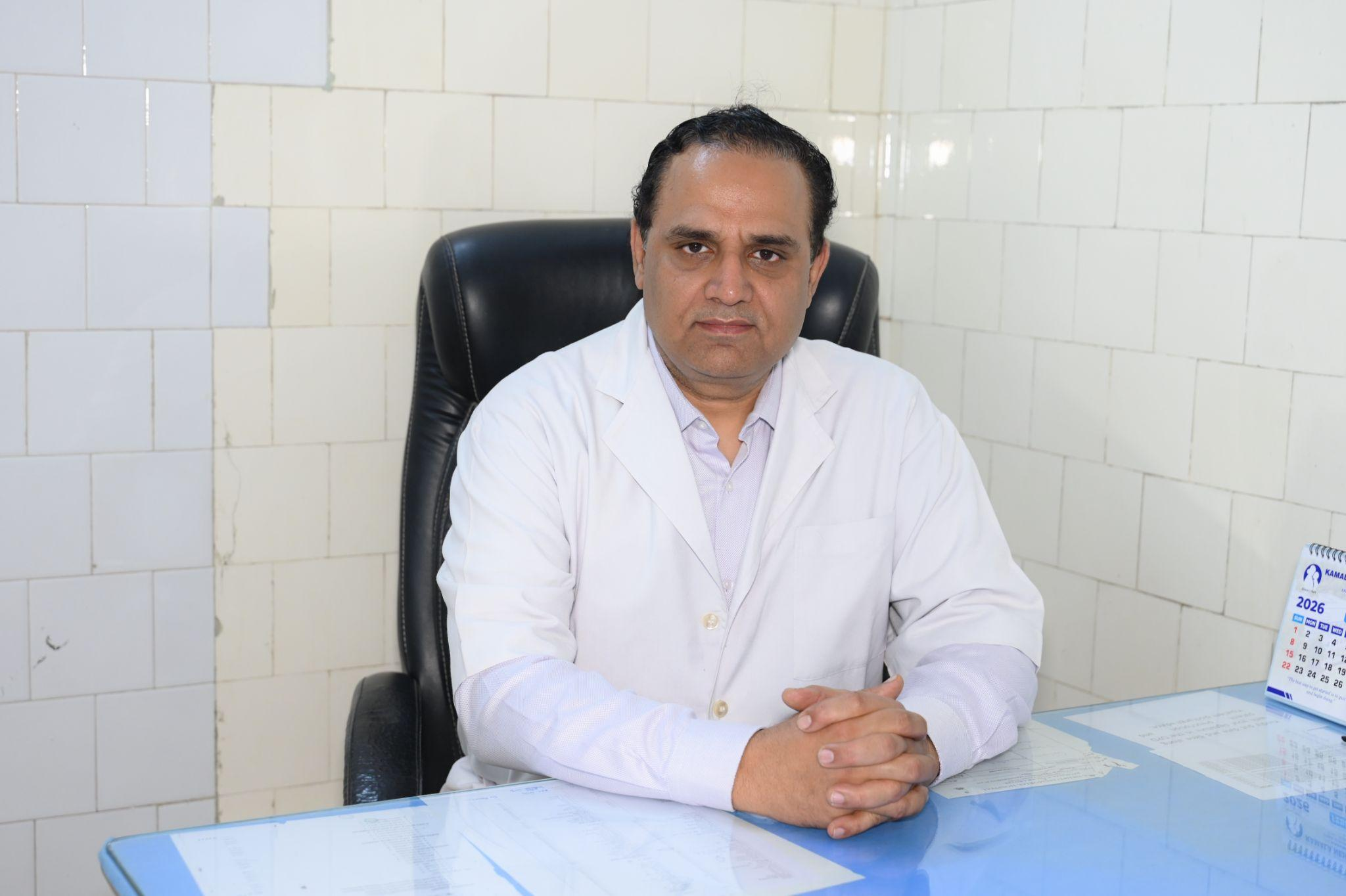Man in a white lab coat sits at a glass desk with folded hands, facing the camera in a tiled office setting.