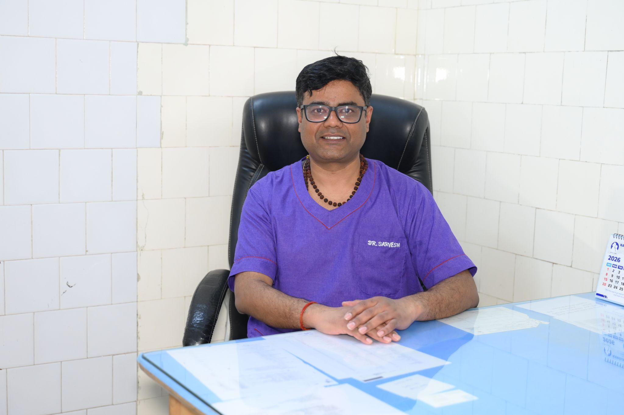 Dr. Sarkesh wearing a purple shirt with a beaded necklace, seated in a leather chair behind a glass desk in a white tiled room.