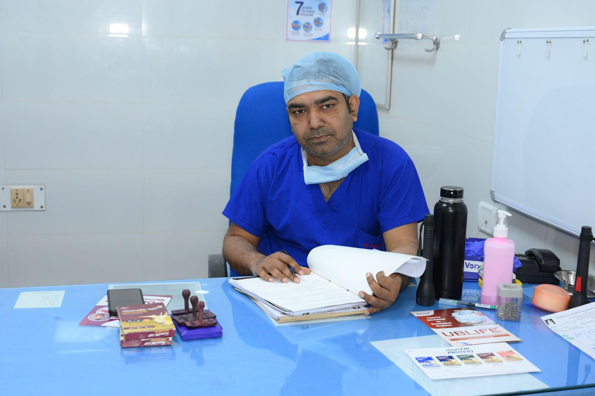 Male medical professional in blue scrubs and cap, reading documents at a blue desk in a clinical room.