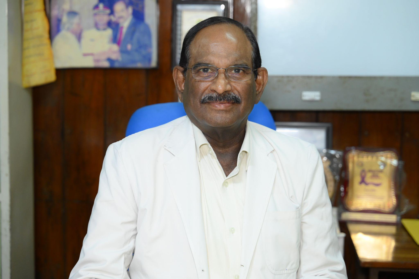 Smiling man wearing a white coat and glasses seated in an office setting with wood-paneled walls behind him.