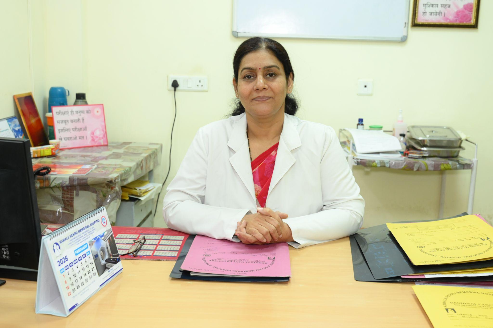 Female doctor seated at a desk in a clinic, hands folded, wearing a white coat over a red outfit, calm expression amid office supplies and charts on the desk.