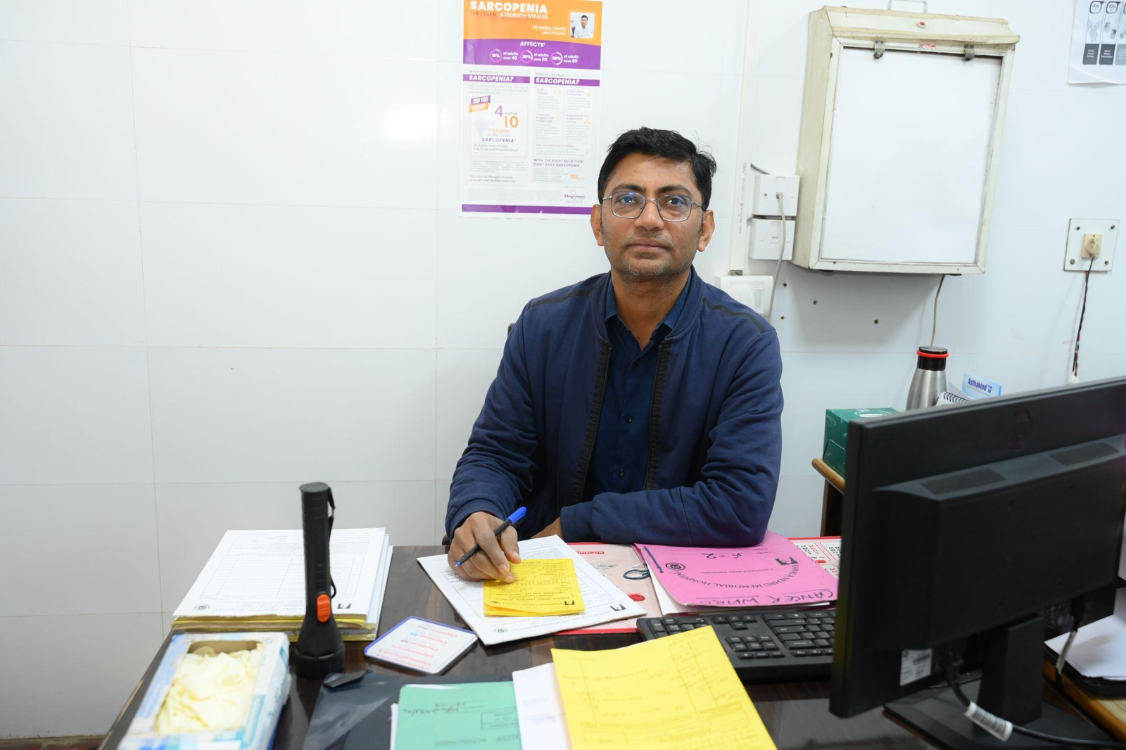 Man in a blue shirt sits at a cluttered desk, writing on papers with a pen, a computer monitor to the right.