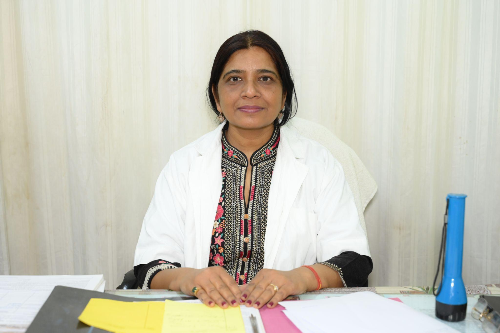 Woman in a white lab coat sits at a desk with colorful printed clothing, facing the camera among scattered papers.