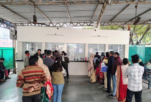 Queue of people at a service desk with glass partitions under a corrugated metal roof; staff assist customers.