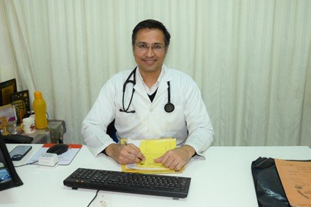 Male doctor in a white coat with a stethoscope sits at a desk, writing on a yellow notepad with a computer keyboard nearby.