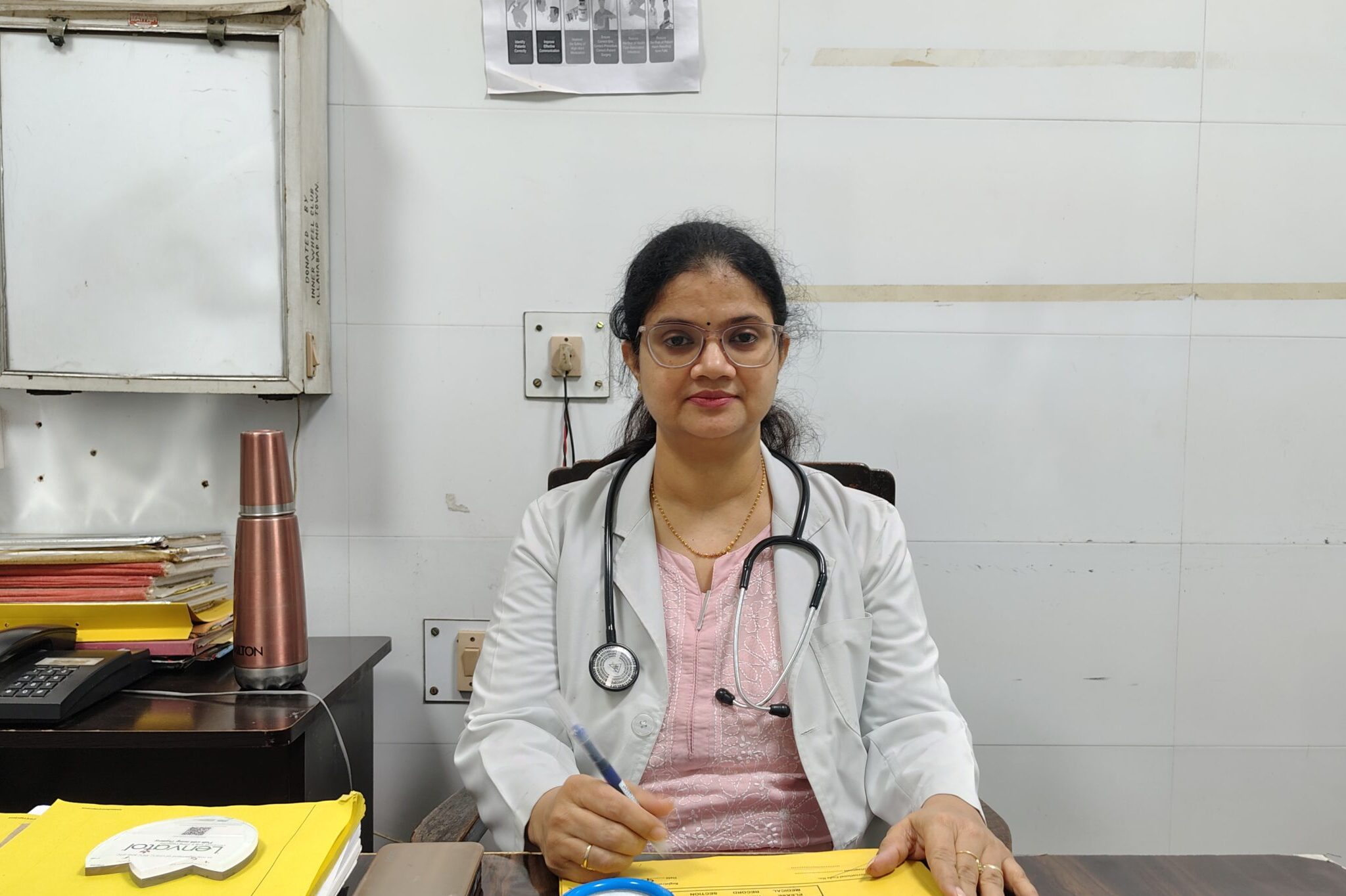 Female doctor in a white coat with a stethoscope around her neck sits at a desk, writing on yellow folders.